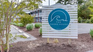 A sign for "The Shore House" with a wave logo, surrounded by landscaping and a building in the background. - The Shore House (Ocean City)
