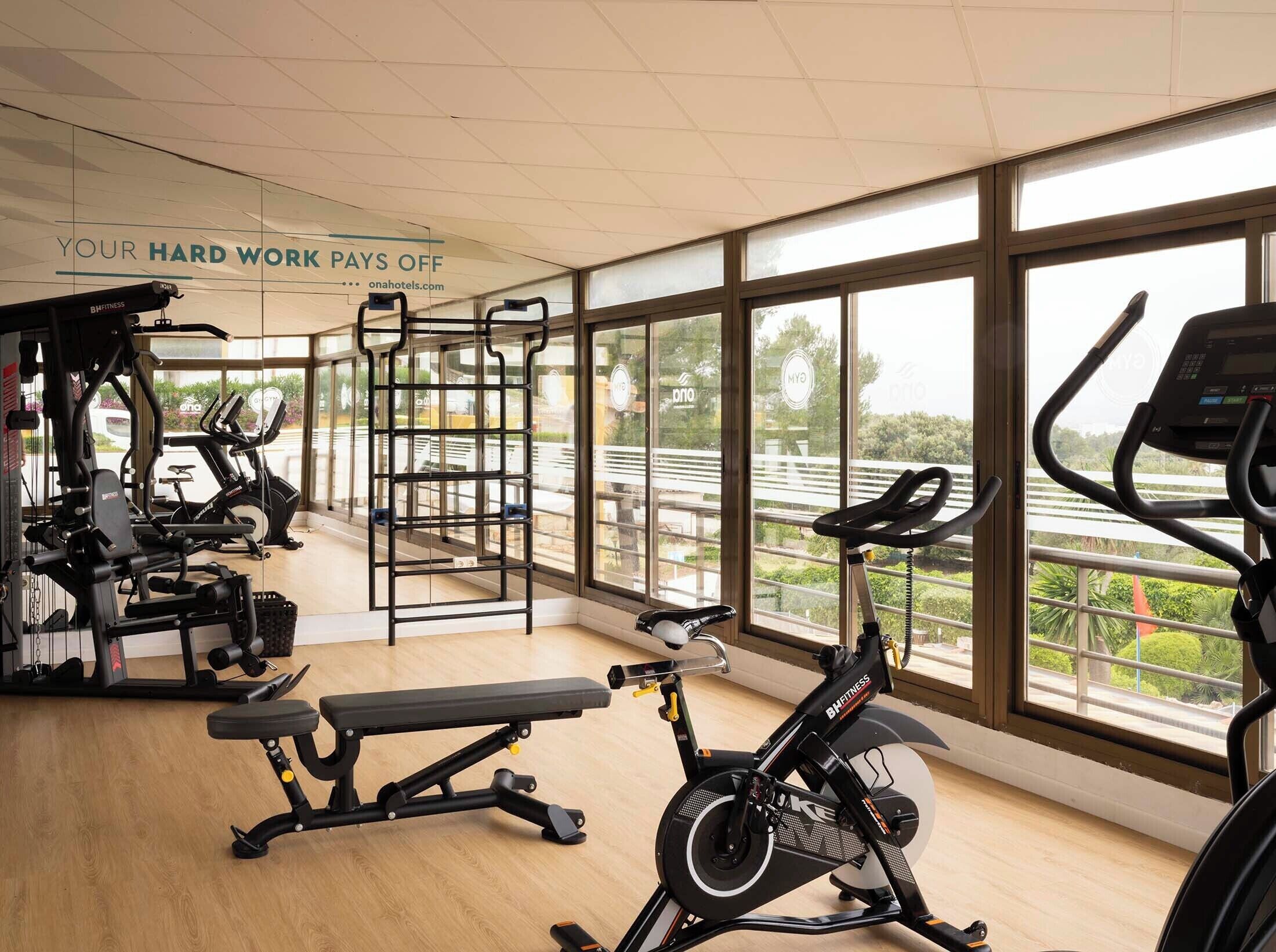 A gym with various exercise equipment, large windows, and a sign stating 'YOUR HARD WORK PAYS OFF'.