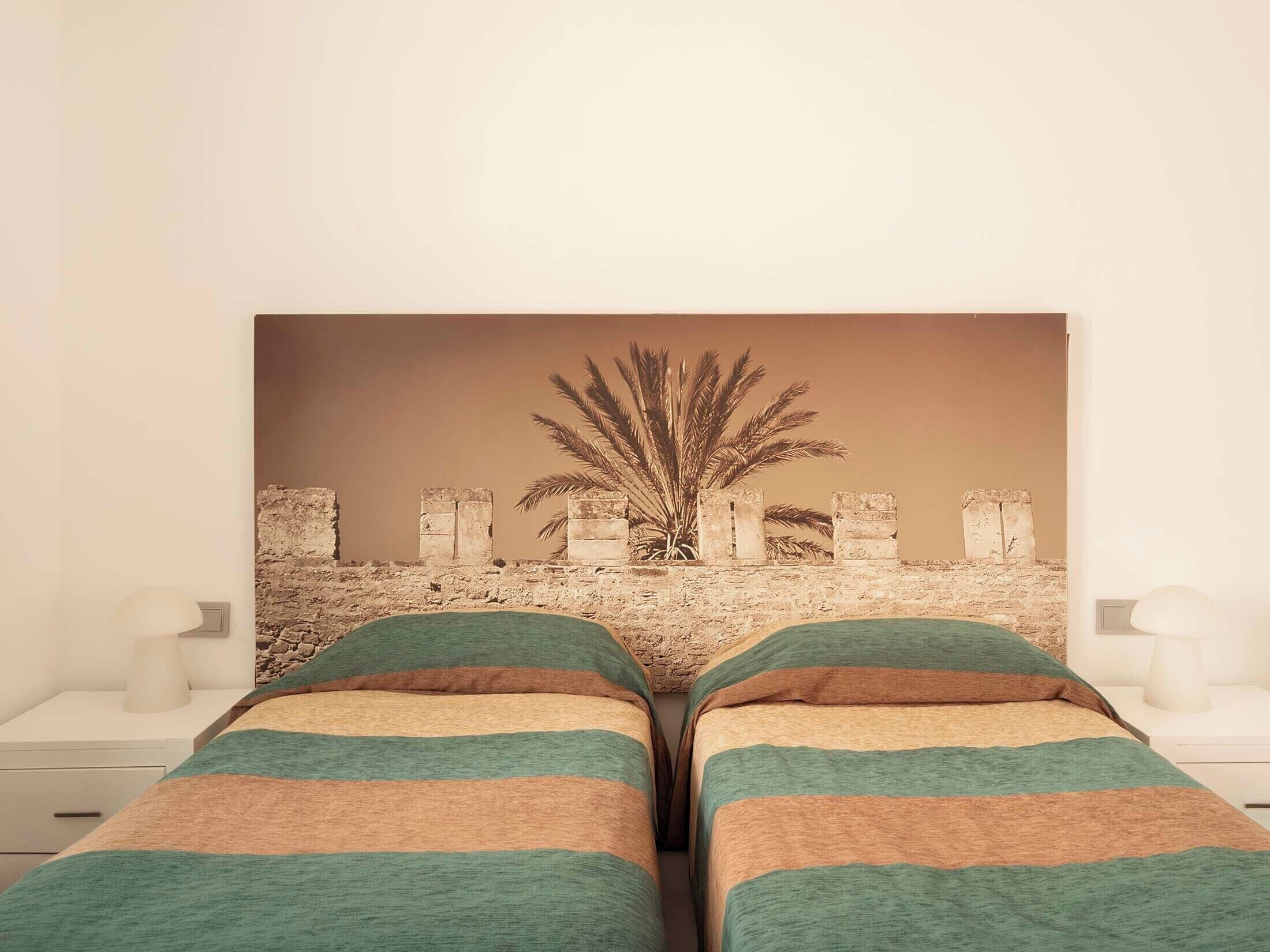 A room with two beds, a wall-mounted artwork of a palm tree, and a stone wall.