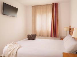 Habitación de hotel con cama, televisión montada en la pared, ventana con cortinas y mesita de noche con lámpara.