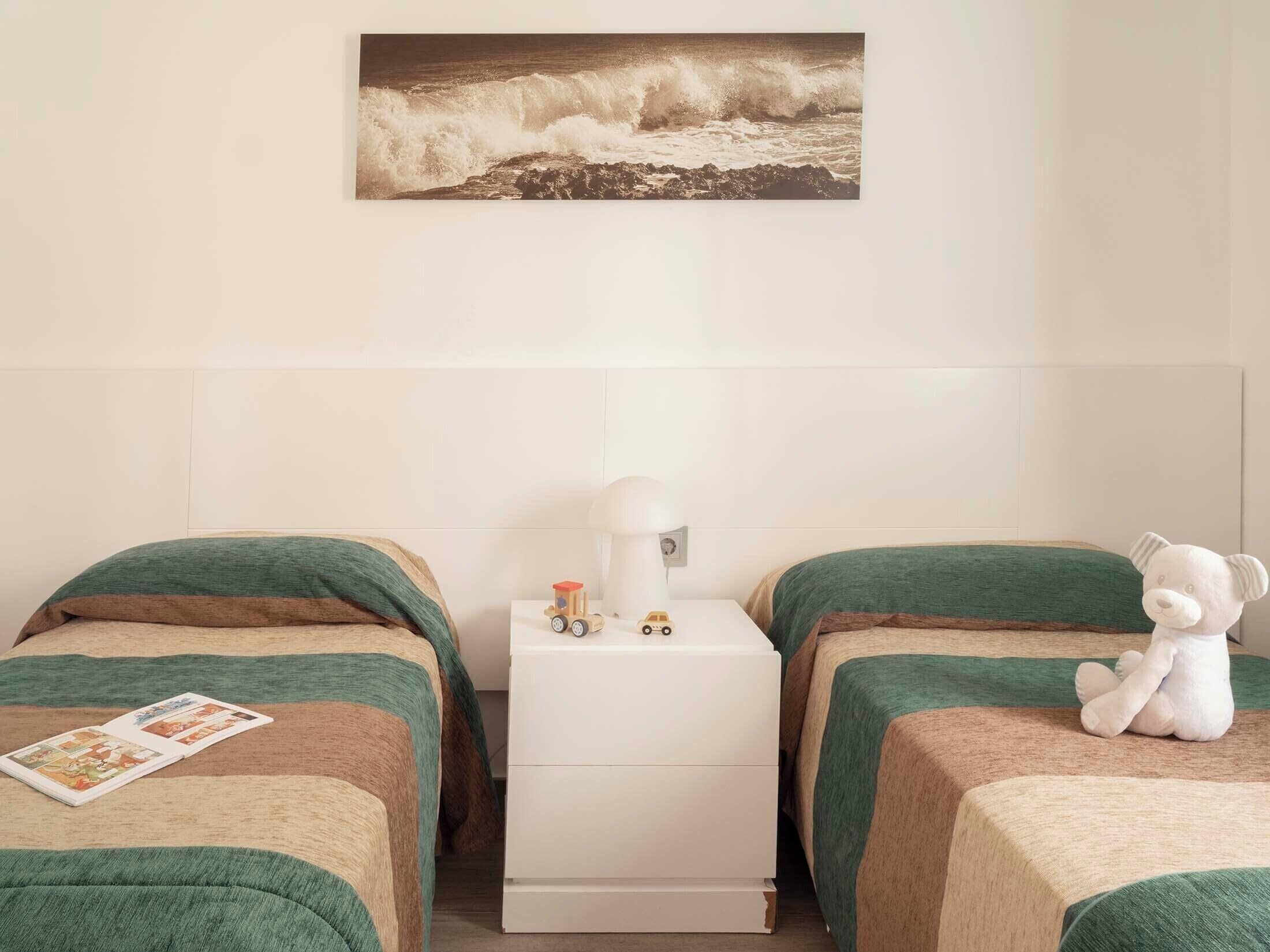 Habitación de hotel con dos camas, una mesita de noche, una lámpara, un oso de peluche y un cuadro con olas en la pared.