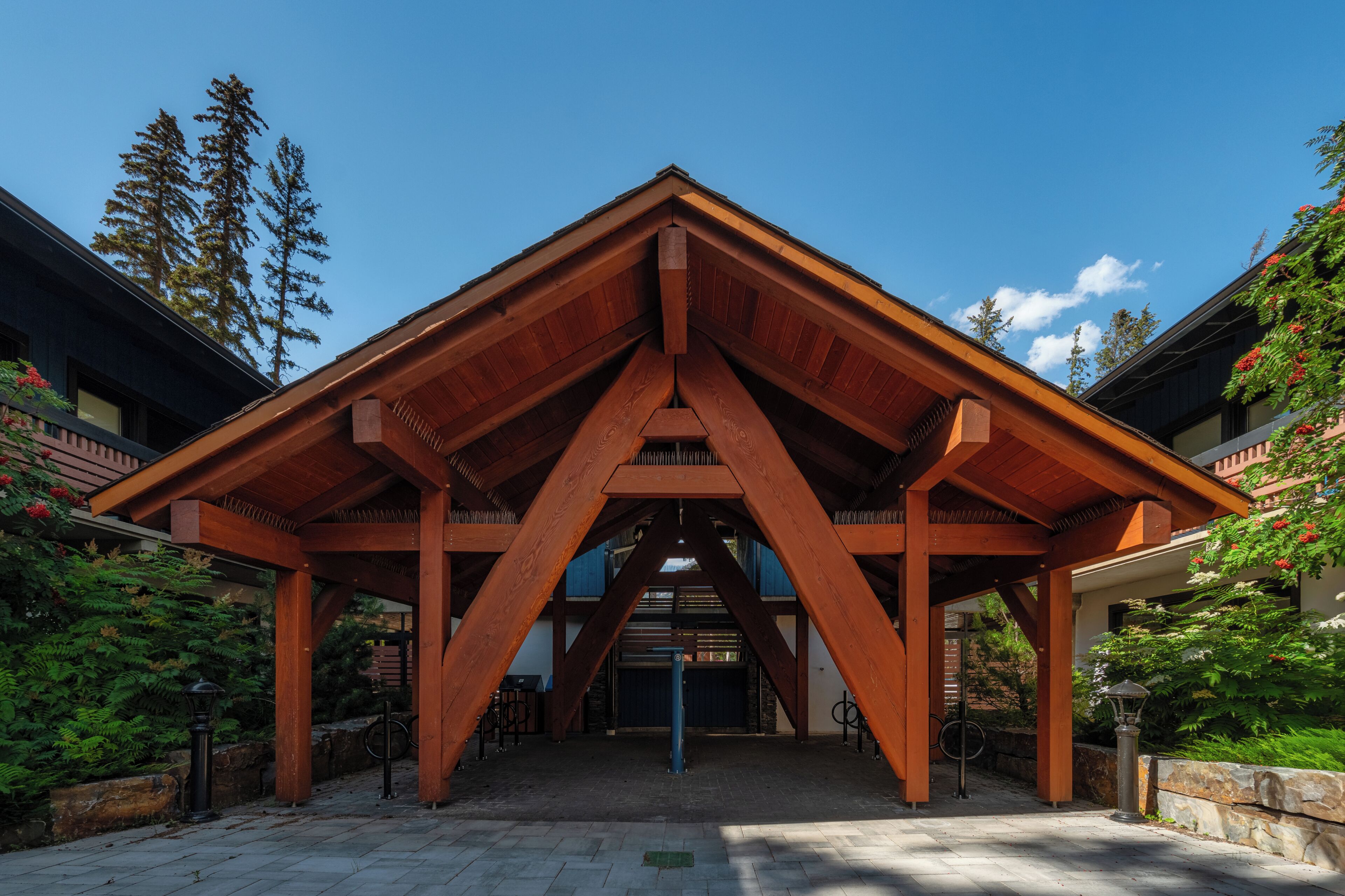 A wooden pavilion with a triangular roof structure, surrounded by greenery and trees.