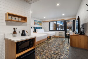A compact hotel room with a kitchenette, a bed, a desk, and a large window offering a view of the outside.