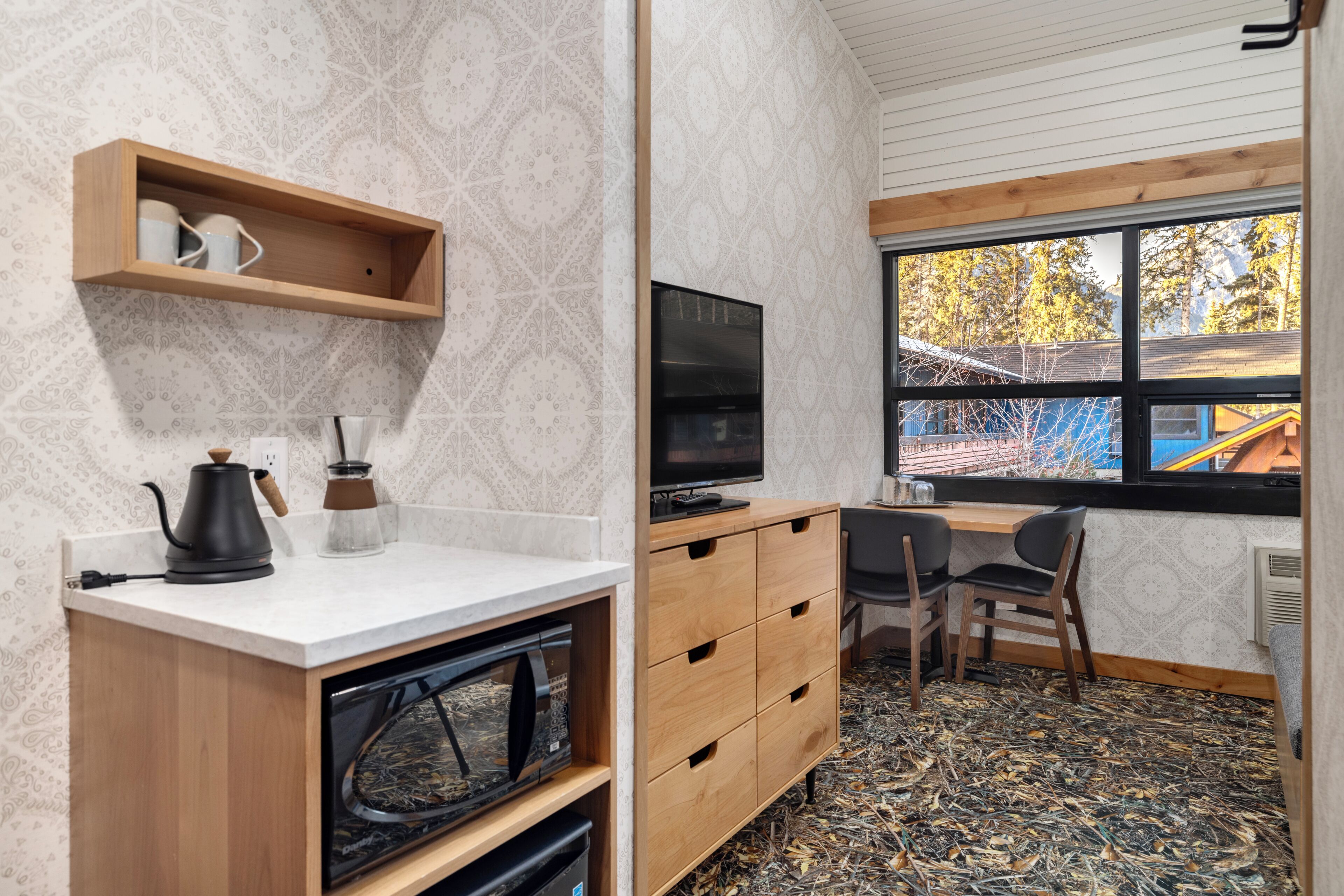 A compact room with a kitchenette, a television, a window with a view of trees, and a wooden desk.