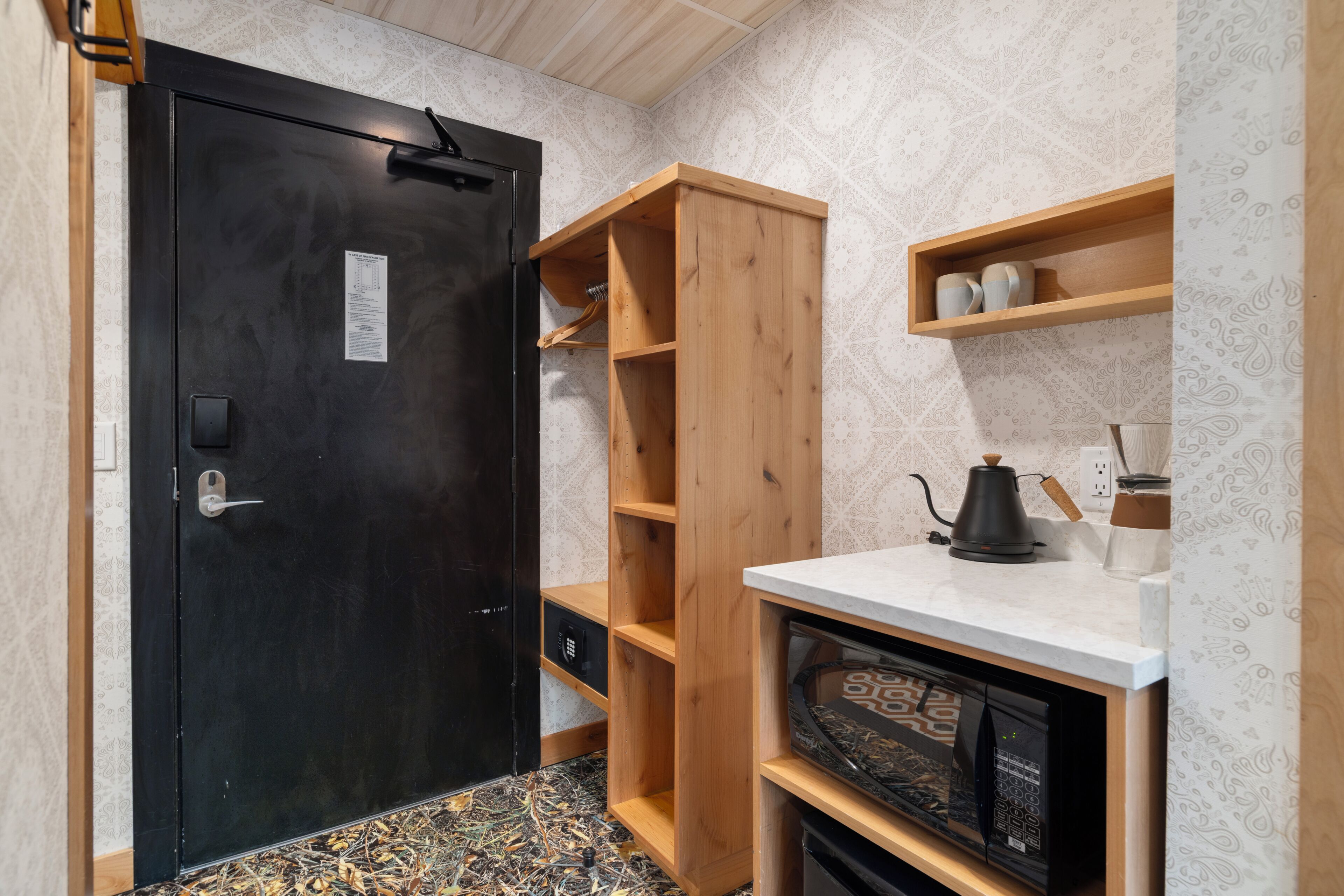 A compact kitchen with a black door, wooden shelves, a microwave, and a coffee maker.