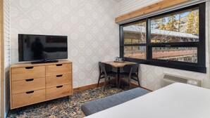 Quarto com televisão em uma cômoda de madeira, uma mesa pequena com cadeiras e janela com vista para árvores e um edifício.