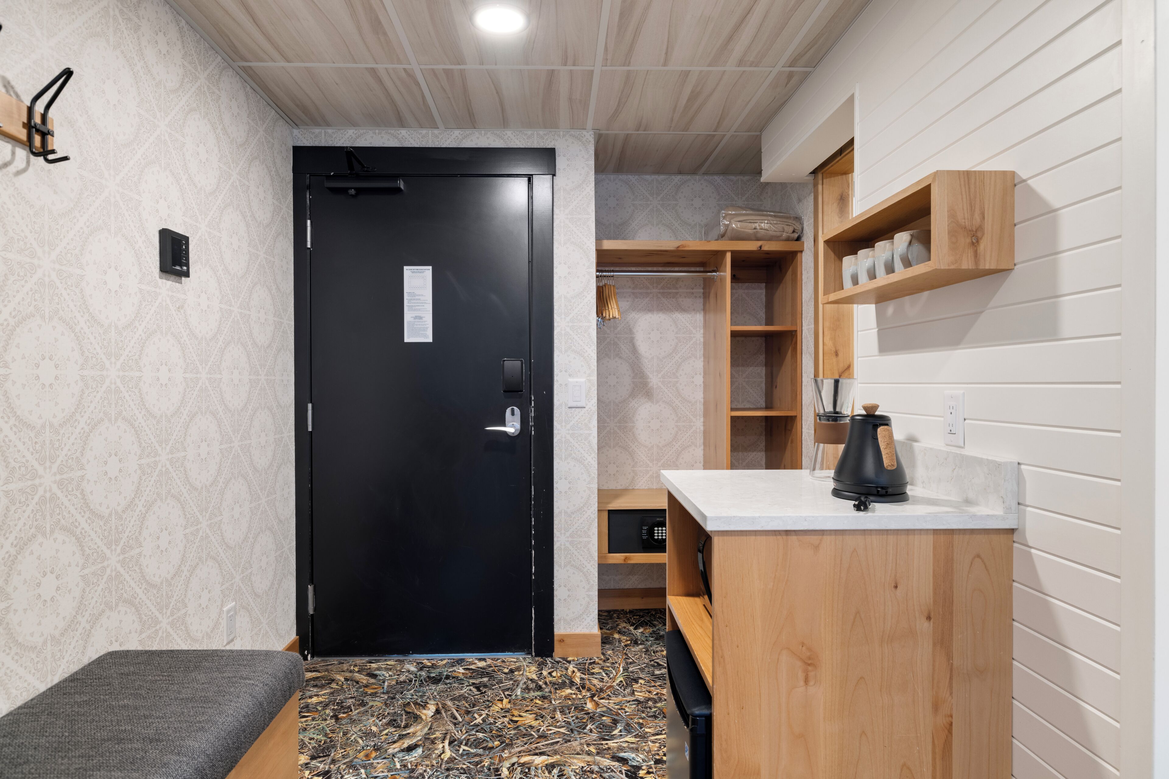 Une pièce de taille modeste avec une porte noire, un petit coin salon, un meuble en bois doté d’étagères et un plan de travail sur lequel se trouve une bouilloire.