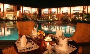 A dining table by a poolside with a candle, wine glasses, and a vase of flowers. - The Golden Crown Hotel & Spa, Colva (Colva)
