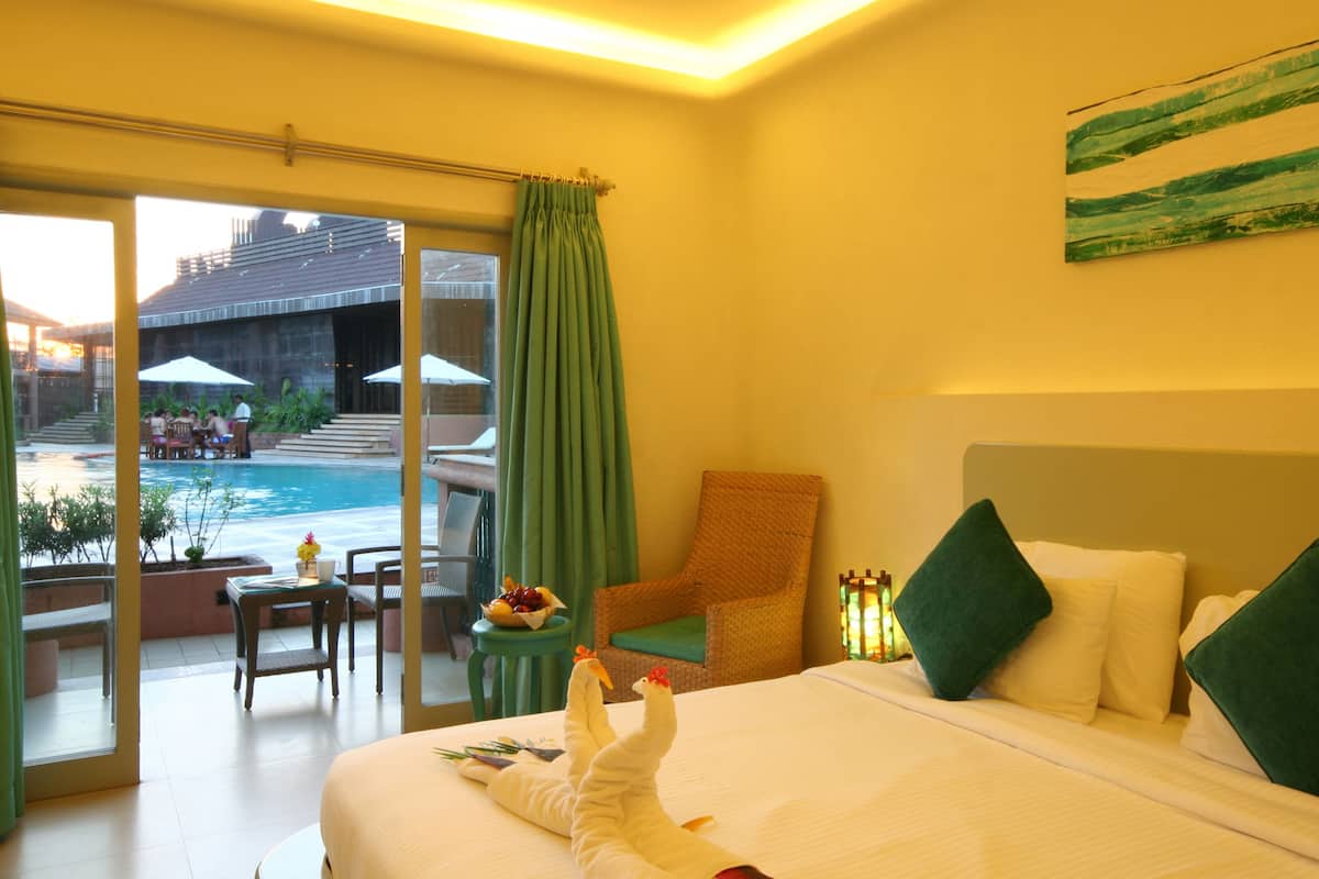 Habitación de hotel con cama, una silla, una mesita con frutero y vista a una piscina al aire libre.