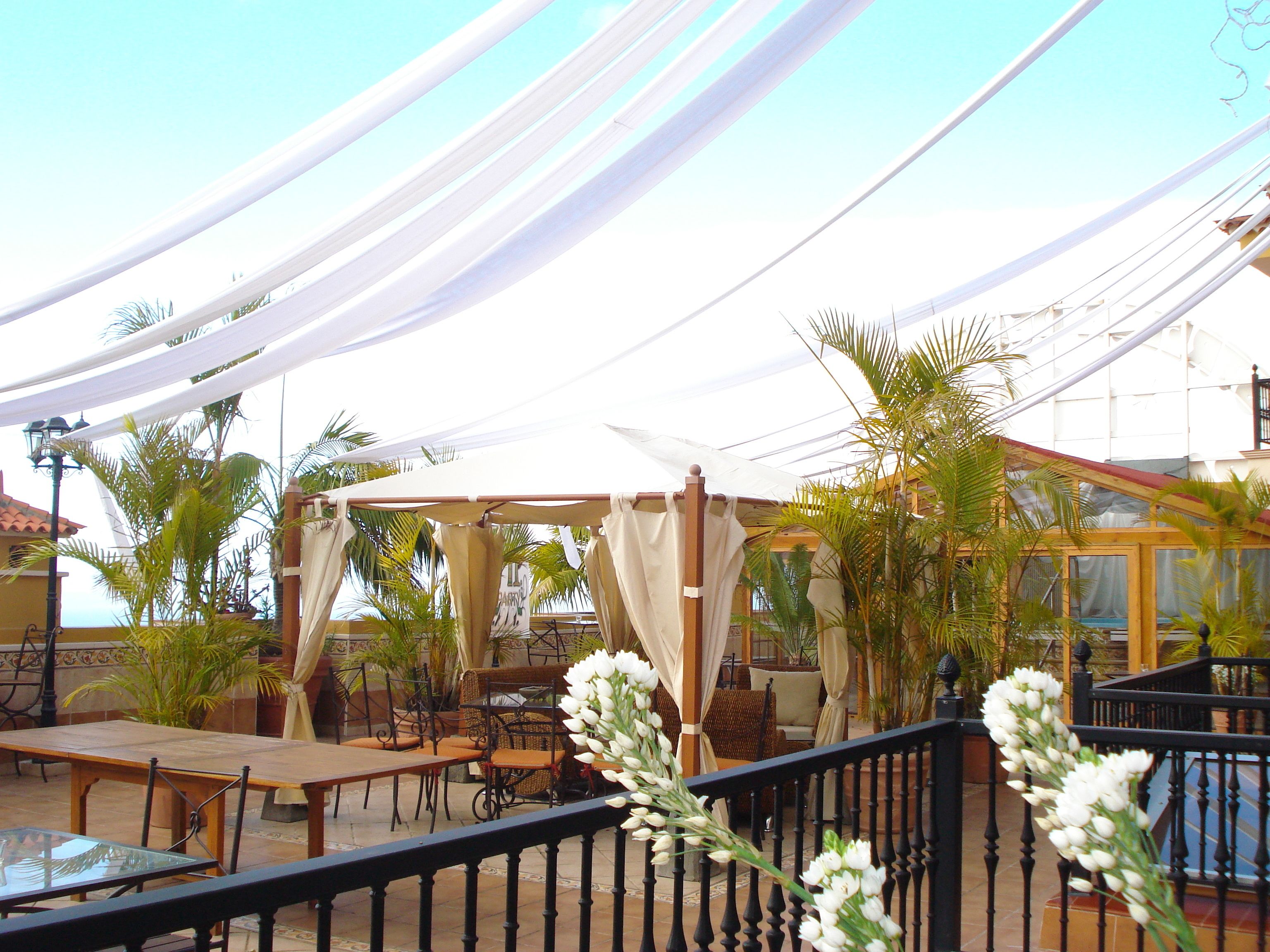 Un espace extérieur aménagé en terrasse, avec des tables et des chaises, un coin salon abrité et une rambarde ornée de fleurs blanches.