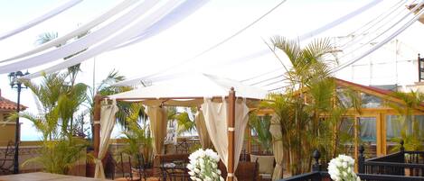 Un espace extérieur aménagé en terrasse, avec des tables et des chaises, un coin salon abrité et une rambarde ornée de fleurs blanches.