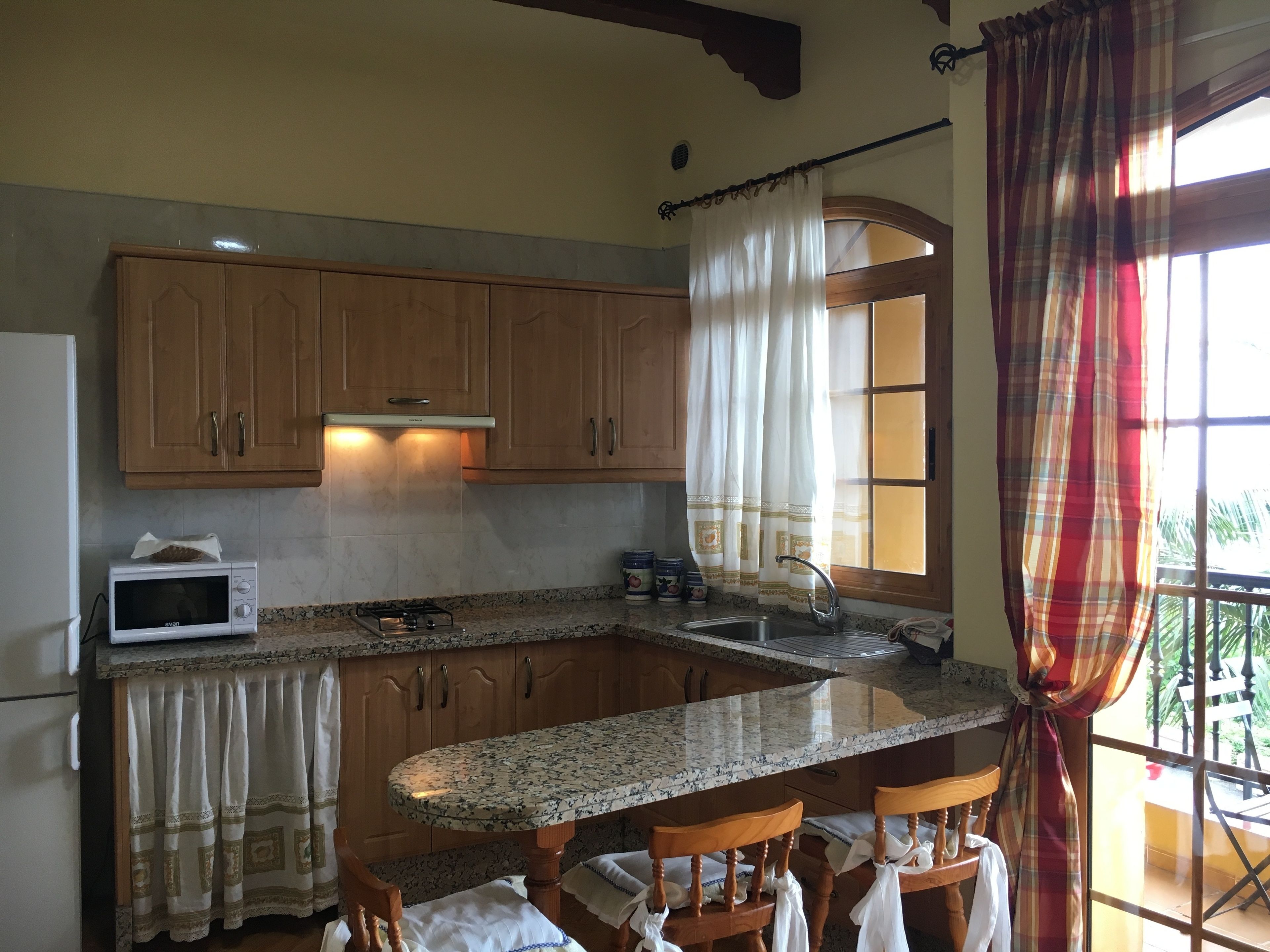 Una cocina con armarios de madera, un microondas, una estufa a gas y una ventana con cortinas.