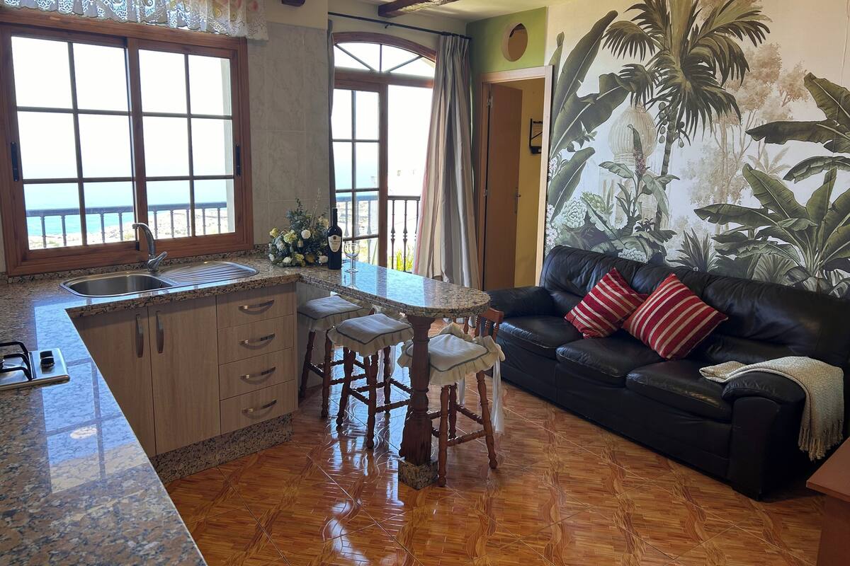 A kitchen with a sink, stove, and dining area with stools. There is a black leather sofa with red and white pillows. A large window provides a view of the ocean.