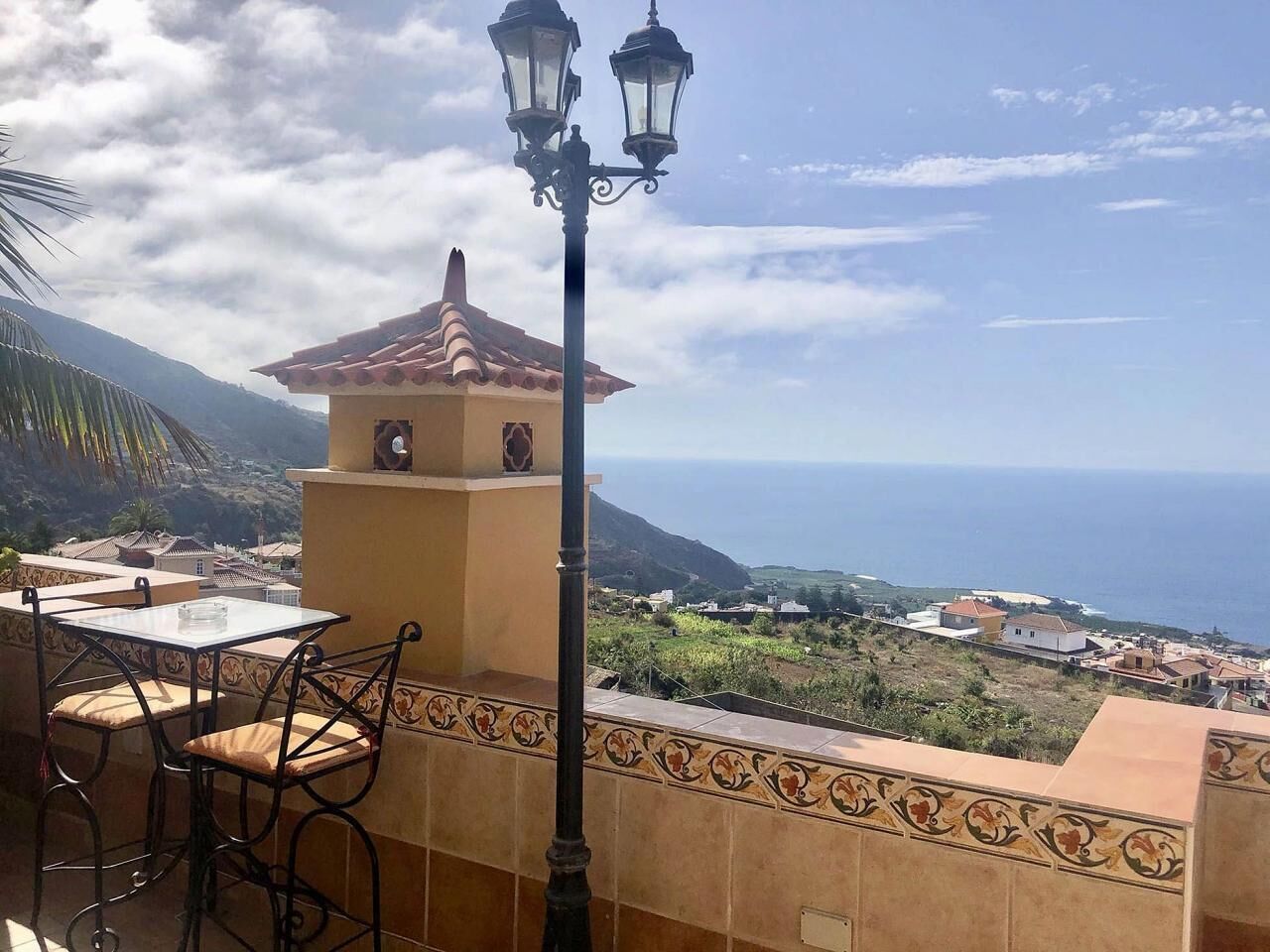 Un balcó amb taula i cadires, una làmpada decorativa i vista al mar i edificis allunyats.