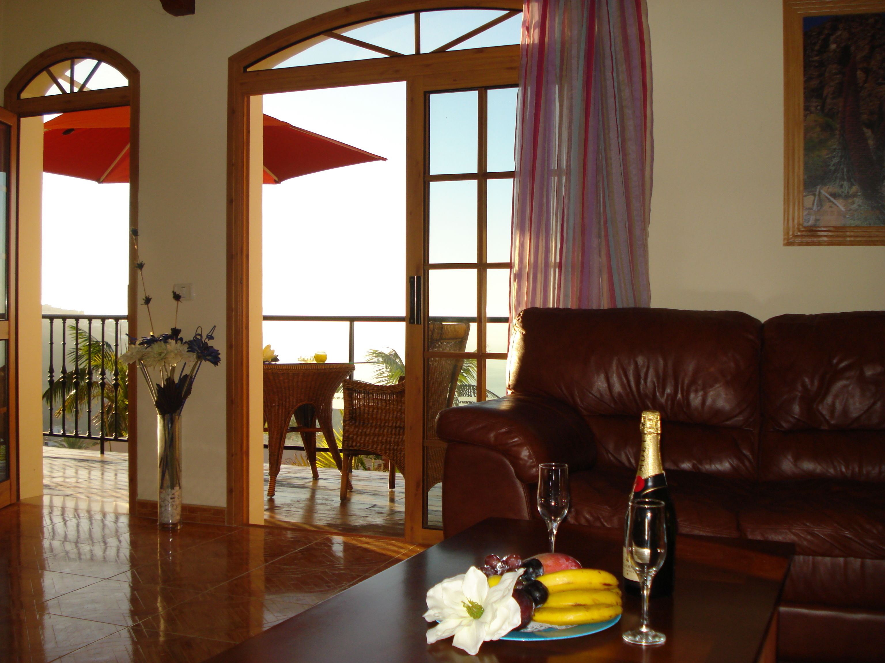 A living room with a leather sofa, a dining table with fruit and a bottle of champagne, and a view of the outdoors through large windows.