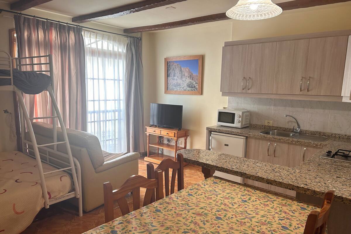 A cozy room with a bunk bed, a dining table with chairs, a kitchen area with a microwave and sink, and a window with curtains.