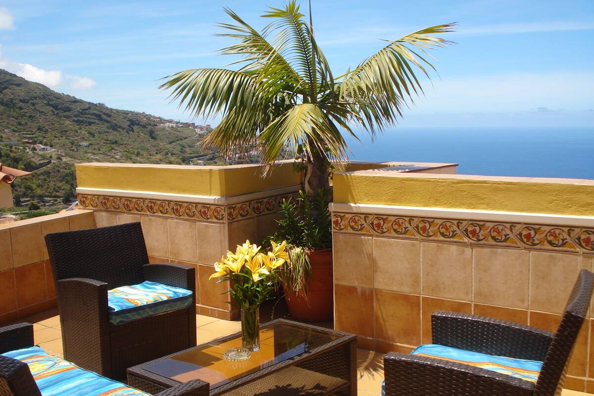 A terrace with wicker furniture, a glass-top table, and a vase of yellow flowers.