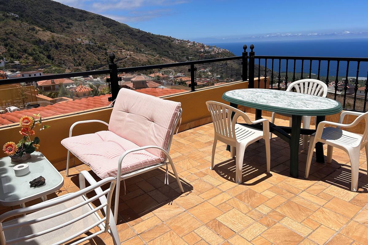 A terrace with a table and chairs, a vase with flowers, and a view of the sea and hills.