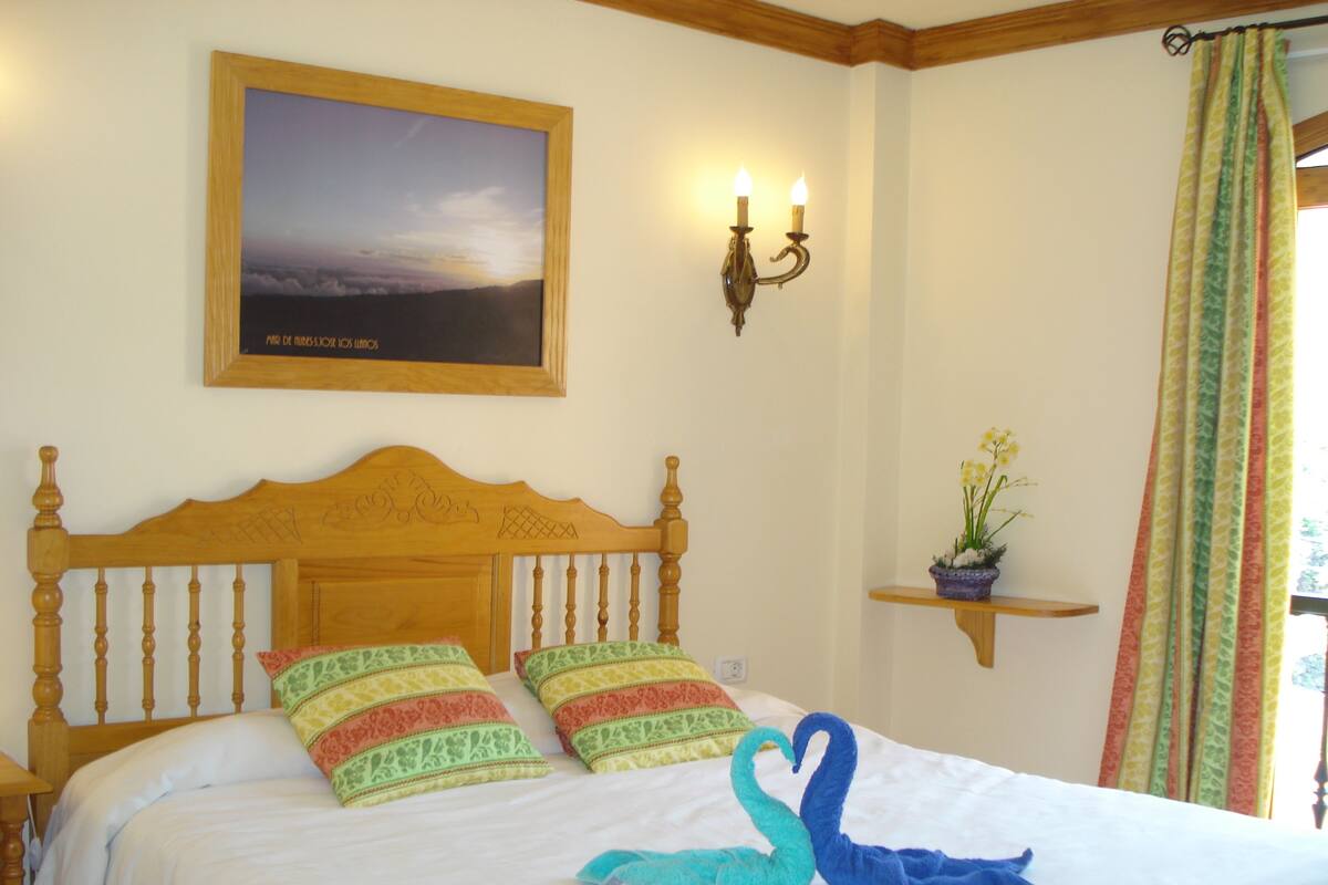 A bedroom with a wooden headboard, two patterned pillows, a swan-shaped towel, a framed picture on the wall, a wall-mounted light fixture, and a window with curtains.