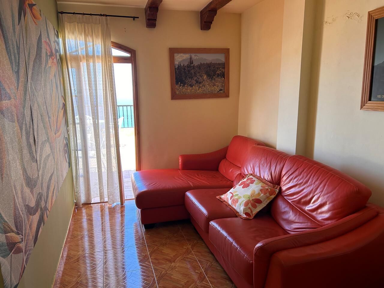Un sofá de cuero rojo con un cojín floral en una habitación con suelo de madera y un cuadro en la pared.