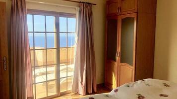 A bedroom with a sliding glass door to a balcony, a wooden wardrobe, and a bed with a floral bedspread.