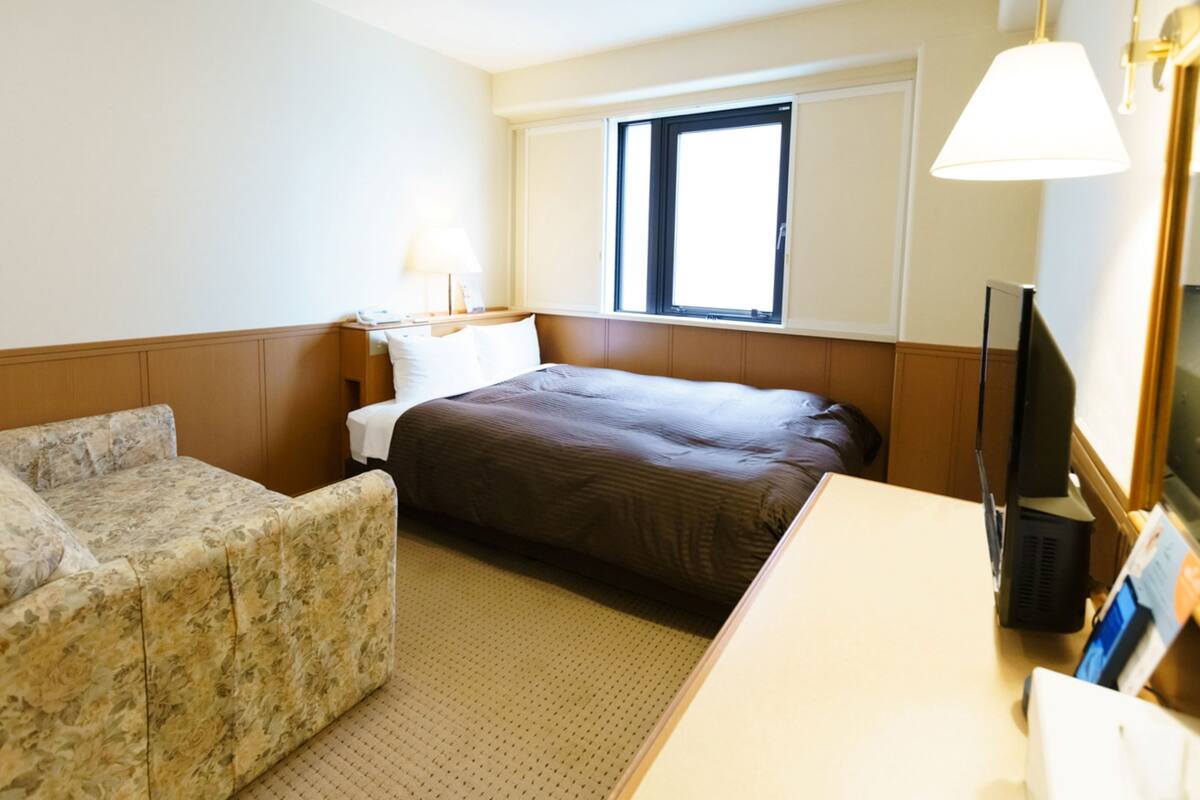 Quarto de hotel com cama, sofá, escrivaninha, televisão e janela com persianas.