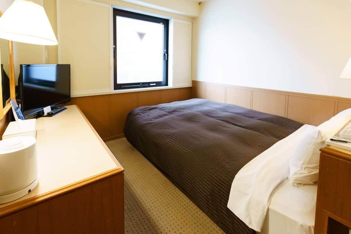 Quarto de hotel com cama, escrivaninha com computador, janela e abajur.