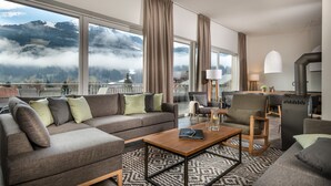 Una sala de estar moderna con un sofá, sillones y una mesa de centro. Amplias ventanas ofrecen vistas a montañas y nubes.
