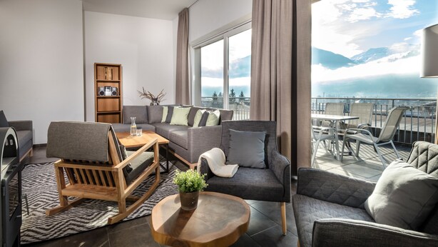 Una sala de estar moderna con un sofá, sillas, una mesa de centro y una estantería. Cuenta con ventanas amplias con vistas a las montañas.