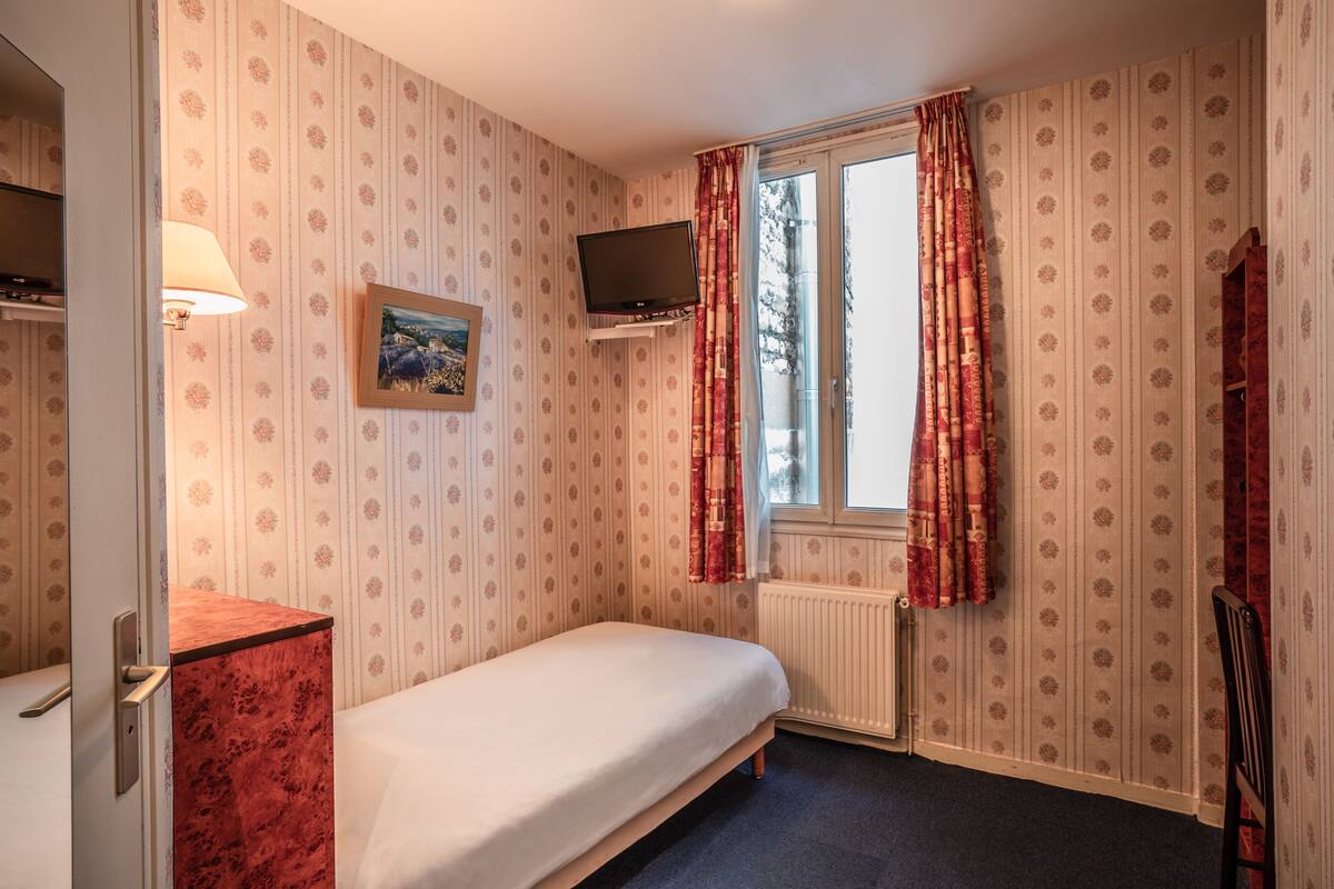 A single bed in a room with striped wallpaper, a mounted television, a window with red curtains, and a radiator.