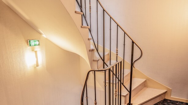 A spiral staircase with a handrail, carpeted steps, and a wall-mounted light fixture. - Hotel Neptune (Paris)