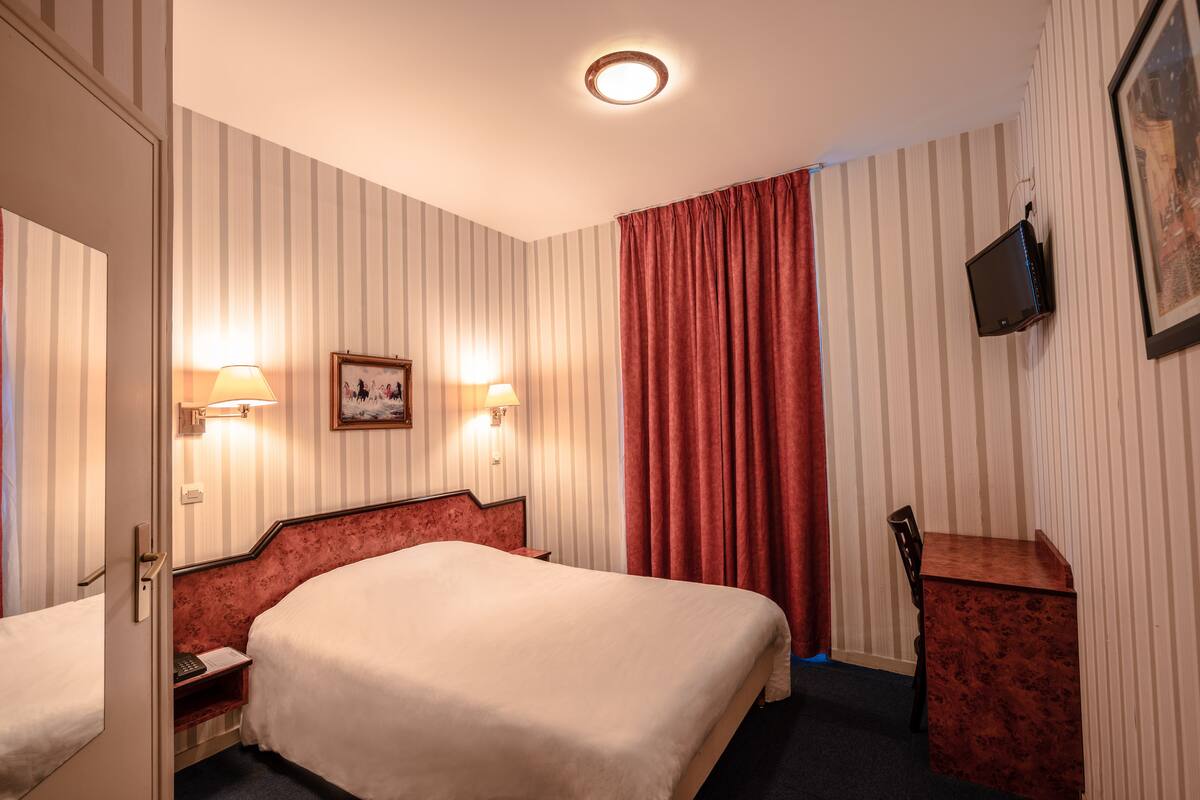 A hotel room with a double bed, bedside lamps, a small desk, a television mounted on the wall, and a window with curtains.
