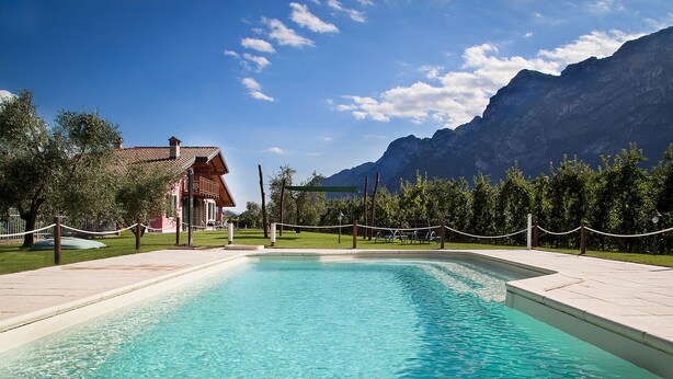A rectangular swimming pool with a tiled deck, surrounded by a rope fence, with mountains in the background. - Agritur La Cort (Arco)