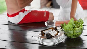 A man and a woman sitting at a wooden table with a pizza oven in the background, a bowl of lettuce, and bottles of beer. - Agritur La Cort (Arco)