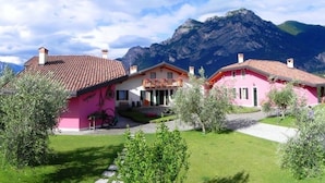A pink house with a tiled roof, surrounded by green grass and trees, with mountains in the background. - Agritur La Cort (Arco)