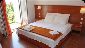 A neatly made bed with white linens and a brown throw blanket, wooden headboard, bedside lamps, and a view of greenery through the window. - Gargamelo Pension (Porec)