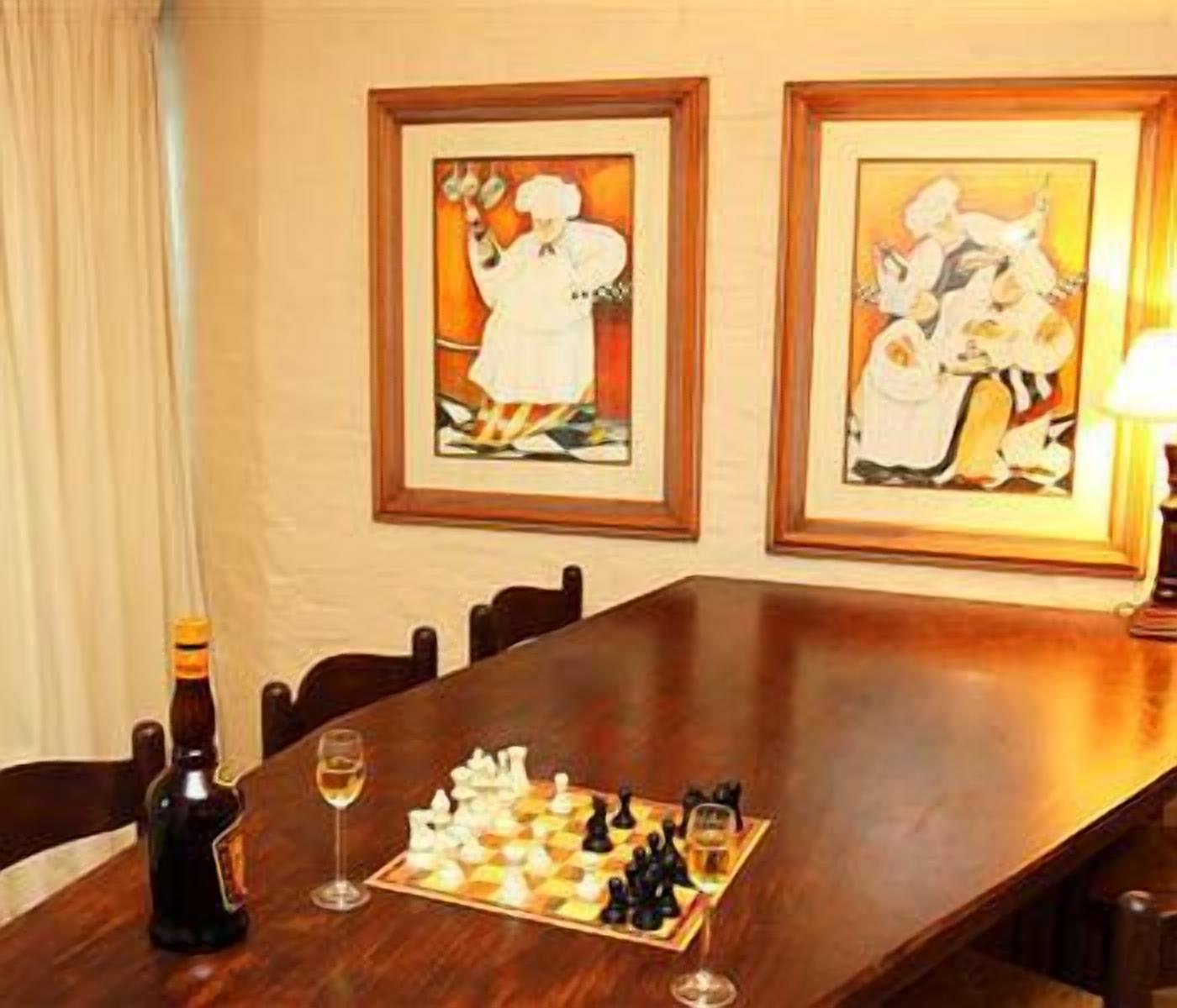 Una mesa de comedor con un juego de ajedrez, copas de vino y una botella. En la pared hay cuadros enmarcados.
