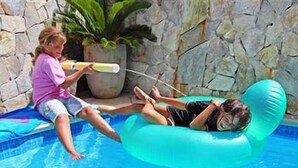 A person lying on a blue inflatable float in a swimming pool with a stone wall and potted plant in the background.