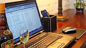 A laptop on a desk with a bottle of wine, a glass of wine, a mouse, and a vase with flowers.