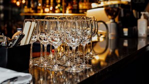 Een bargedeelte met meerdere glazen op een toonbank, een fles alcohol en een plank met diverse flessen op de achtergrond.