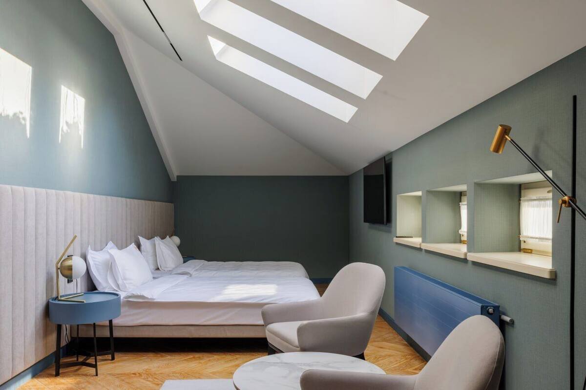 Un dormitorio moderno con techo inclinado, tragaluces, cama, sillón, mesita de noche y televisor montado en la pared.