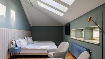 A modern bedroom with a sloped ceiling, skylights, a bed, a chair, a bedside table, and a wall-mounted TV.