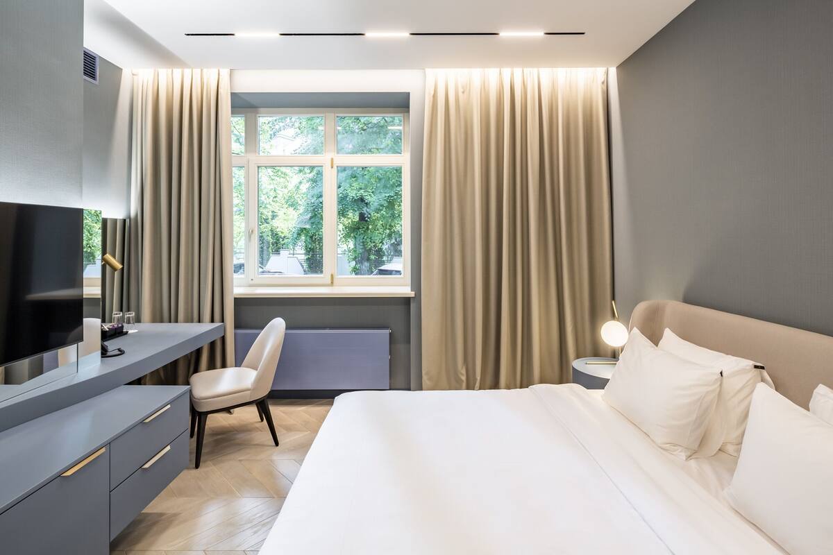 Una habitación de hotel moderna con cama, escritorio con televisor, silla y ventana con cortinas.