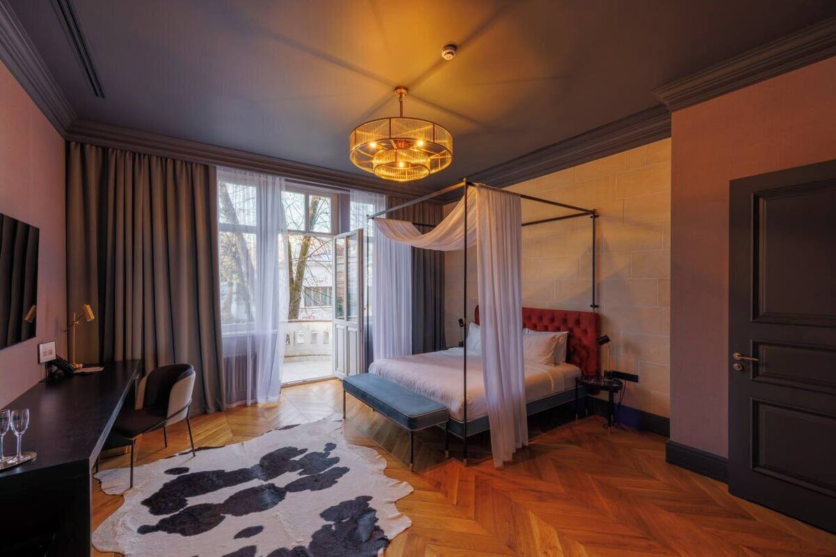 Un dormitorio con cama con dosel, un escritorio con silla, un ventanal con cortinas y un candelabro.
