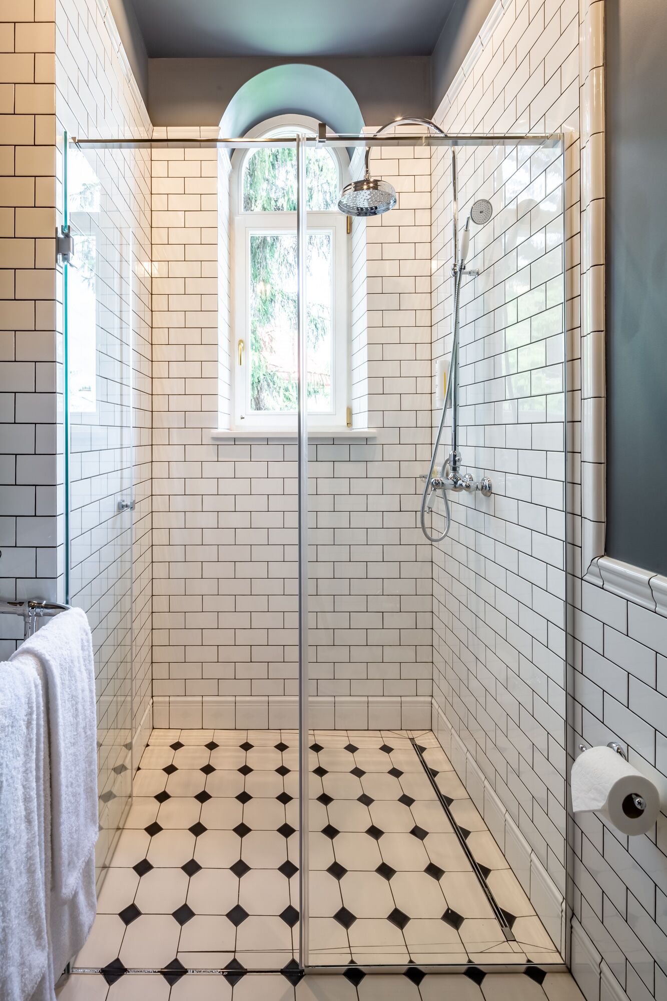 A walk-in shower with white and black patterned tiles, a glass door, and a window with a view of greenery.