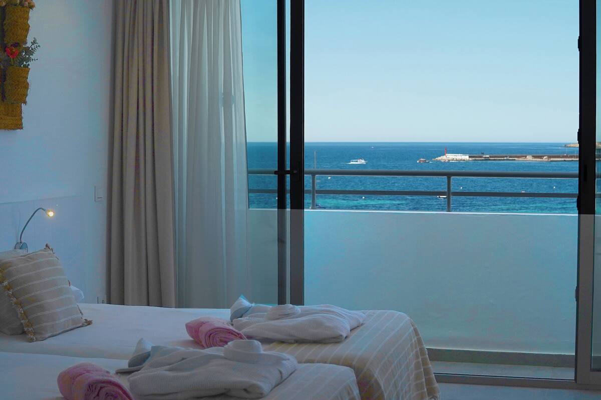 Een hotelkamer met een bed, een nachtkastje en uitzicht op zee door grote ramen.