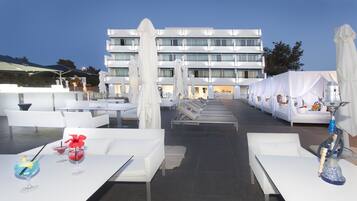 Una terraza en la azotea con sillones blancos y mesas, un edificio grande al fondo y un narguile a la derecha.