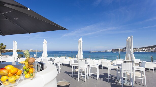 A seaside terrace with white wicker furniture, a fruit bowl, and umbrellas. - Marina Playa Hotel Apartments by Typic Hotels – Adults Only (Sant Antoni de Portmany)