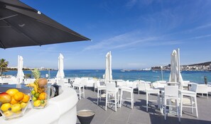 Eine Terrassenanlage am Meer mit weißen Korbmöbeln, einer Obstschale und Sonnenschirmen.