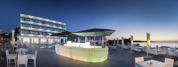 Un hotel frente al agua con terraza, piscina y zona de bar.